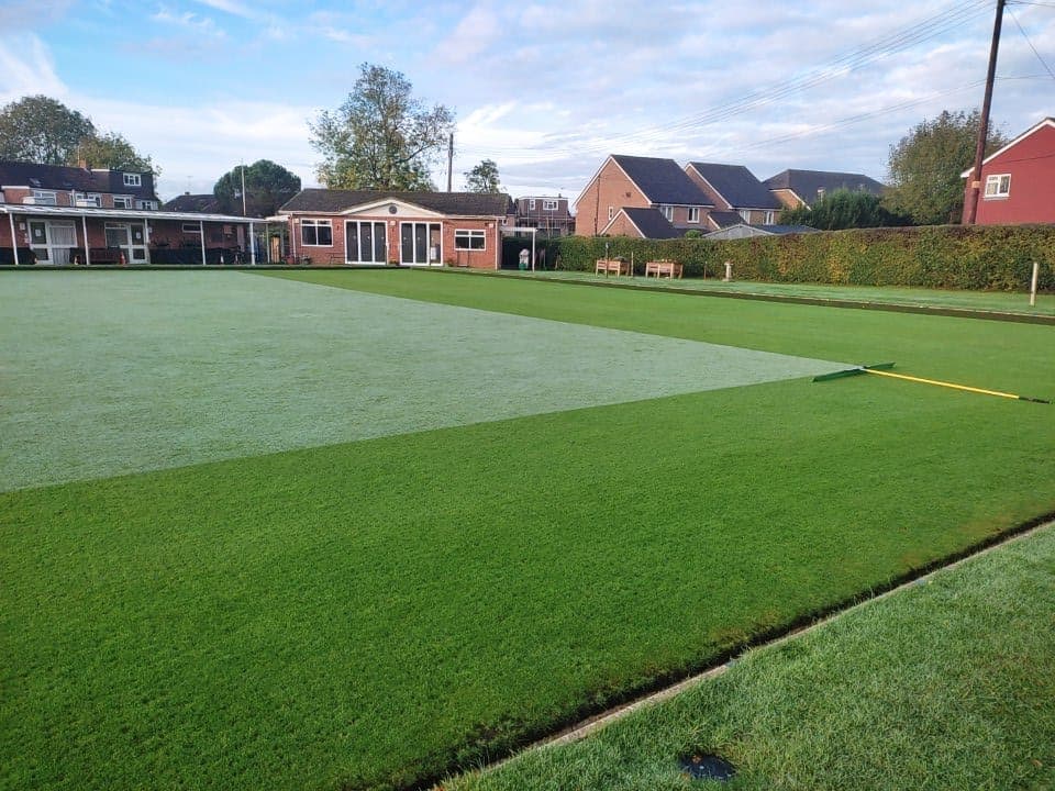 The bowling green half swept — the darker swept half has dew brushed in and grass visible, the lighter silvery half still has its dew coating